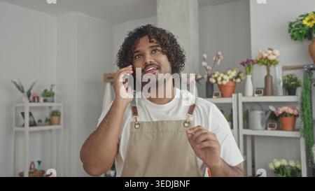 Mann, der mit Handgesten auf dem Smartphone spricht, lächelnd, Florist trägt Schürze zwischen Blumenregalen in einem Gebäude; Service ruhig. Stockfoto