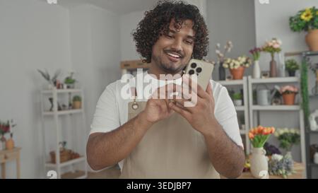 Mann Florist klopft Smartphone mit Händen in einem Blumenladen Gebäude, trägt eine Schürze und lächelt auf den Bildschirm; kleine Unternehmen kümmern sich gut. Stockfoto