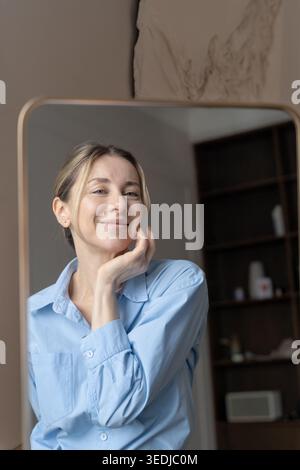 Selbstbewusste zufriedene Frau mittleren Alters steht vor dem Spiegel und schaut auf Reflexion. Eine fröhliche Midlife-Frau lächelt leicht, um Spiegelapplikation zu reflektieren Stockfoto
