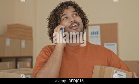 Mann mit Pappkarton und Smartphone, Hand an Ohr beim Lächeln und Reden, umgeben von Paketen im Gebäude; Optimismus für kleine Unternehmen. Stockfoto
