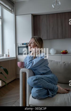 Positive, fröhliche Frau mittleren Alters, die bequem drinnen auf dem Sofa sitzt und mit aufrichtigem Lächeln aus dem Fenster blickt. Reife Frau erinnert sich an etwas Plädoyer Stockfoto