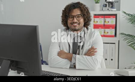 Mann Doktor mit Stethoskop, Arme überkreuzt am Klinikpult, lächelt in die Kamera, Mitgefühl Selbstvertrauen. Stockfoto