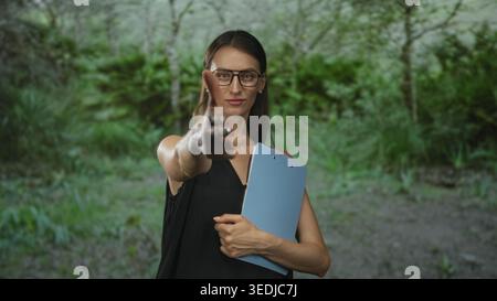 Frau, die eine Brille trägt, mit den Händen blaues Klemmbrett hält, zeigt den Finger nach vorne im Wald; Selbstvertrauen. Stockfoto
