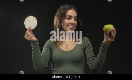 Frau mit grünem Apfel und Reiskuchen, lächelnd, während sie die Entscheidungen im Studio vergleicht; gesunder Optimismus. Stockfoto