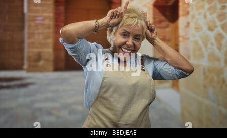 Eine ranghanische Frau mit Schürze zeigt Zeigefinger auf den Kopf in verspielter Horngeste auf der Straße; verspielte Freude. Stockfoto