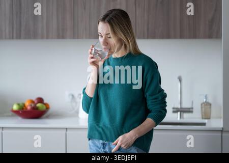 Eine ruhige Frau steht in der Küche verloren in Reflexion, während sie Wasser trinkt. Morgenstimmung der nachdenklichen Frau Stockfoto
