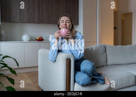 Eine ruhige, zufriedene Frau genießt das Aroma von Heißgetränk mit geschlossenen Augen, Selbstpflegemoment auf der Heimcouch Stockfoto