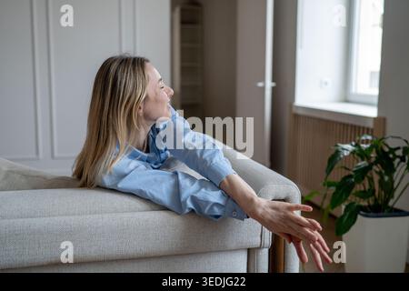 Fröhliche, entspannte Frau, die sich die Arme zum Vergnügen dehnt und sich zu Hause auf dem Sofa ausruhen, fühlt sich wohl. Stockfoto