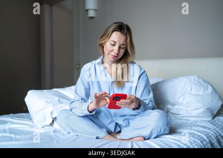 Morgens mit dem Telefon einer Frau, die ihr Smartphone benutzt und nach dem Aufwachen Nachtwäsche trägt. Stockfoto