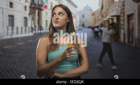 Die junge hispanische Brünette hält dekorierte Eiskegel in der Hand und blickt nach oben, während sie auf einer engen Kopfsteinpflasterstraße mit historischem bui steht Stockfoto