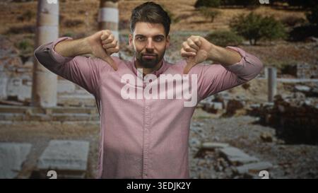 Hispanischer Mann mit Bart zeigt Daumen mit beiden Händen vor dem alten Gebäude aus Steinruinen; Kritik missbilligt. Stockfoto