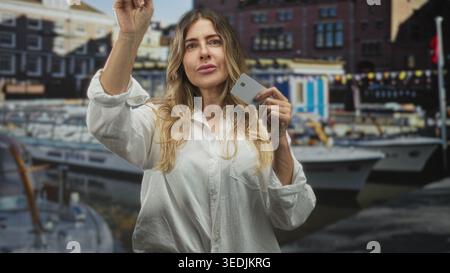 Frau, die eine Kreditkarte hält und mit dem Finger nach vorne zeigt, mit Booten am Hafen; Selbstvertrauen stärken. Stockfoto