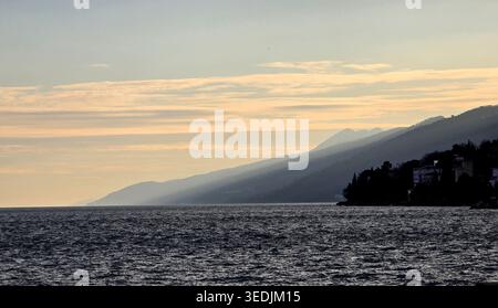 Starke Sonnenstrahlen brechen durch einen trüben Abendhimmel über einem dunklen, plätschernden Meer. Die mehrschichtigen Silhouetten der Berge bilden eine nebelige Horizontlinie entlang einer rauen Landschaft Stockfoto