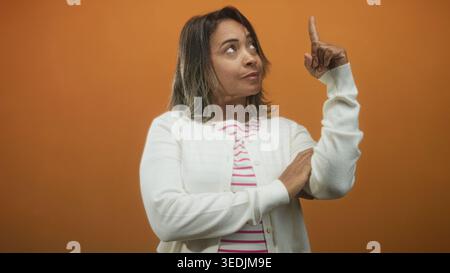 Die Frau zeigt den Zeigefinger nach oben, trägt im Studio eine weiße Strickjacke und ein gestreiftes Oberteil. Stockfoto