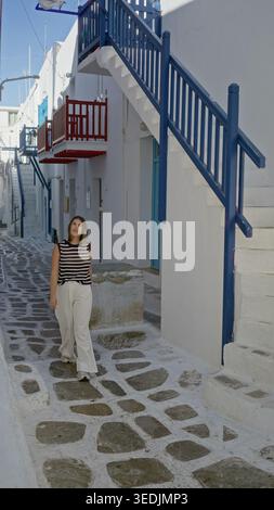 Frau, die mit Hand an Haar an weißen Stufen, blauen Geländern und rotem Balkon in einer engen mykonos Straße vorbeigeht; ruhige Reisegedächtnis. Stockfoto