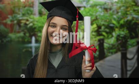 Teenager-Mädchen mit Mütze und Kleid hält die Diplomrolle mit der Hand im grünen Wald; stolz auf die Leistung. Stockfoto