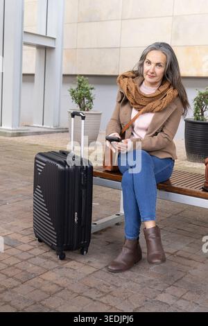 Reife Frau mit Mantel und Schal, die auf der Bank am plaza sitzt und das Smartphone neben dem Koffer hält Stockfoto
