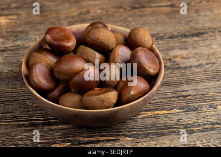 Braune Kastanien in einer Schüssel, reife essbare Kastanien mit glänzenden Fellen liegen in einer Holzschale auf der Oberfläche eines alten dunklen Tisches. Stockfoto