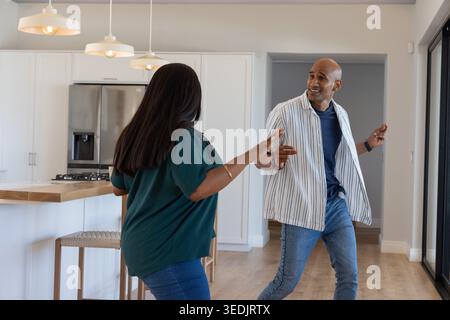 Afroamerikanisches Paar tanzt spielerisch in der hellen Küche nahe der Holzinsel und Pendelleuchten Stockfoto