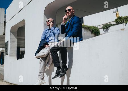 Zwei Geschäftsleute sitzen an einer weißen Wand und unterhalten sich bei Tageslicht in der Stadt auf Mobiltelefonen Stockfoto
