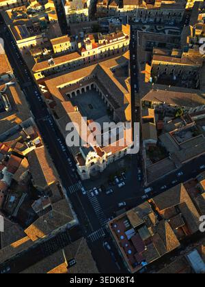Blick aus der Vogelperspektive auf ein historisches Kloster mit einem zentralen Innenhof und bogenförmigen Kreuzgängen, umgeben von Terrakotta-Dächern während der goldenen Stunde in Catania, SIC Stockfoto