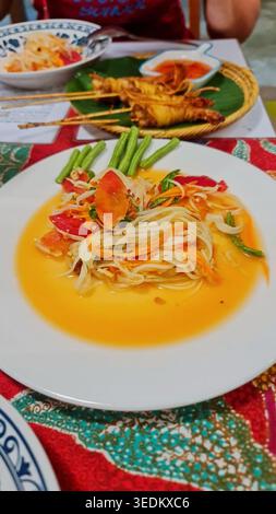 Nahaufnahme mit scharfem thailändischem grünem Papaya-Salat mit Tomaten, langen Bohnen, Chili-Dressing und gegrillten Hähnchenspießen auf einem Restauranttisch. Stockfoto