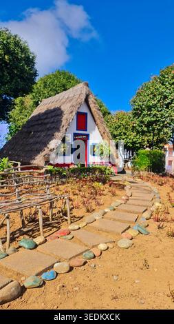 Buntes traditionelles strohgedecktes Ferienhaus mit Steinweg, rustikalem Zaun und üppigen Bäumen unter einem hellblauen Himmel in einem bezaubernden ländlichen Garten. Stockfoto