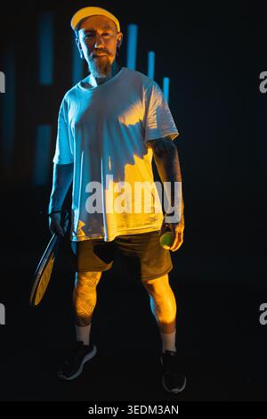 Ein Mann, der einen Tennisschläger und Ball in einem dunklen Raum hält, mit farbigen Lichtern, die auf ihn leuchten Stockfoto