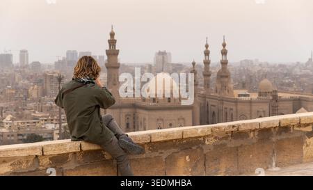 Ein Tourist, der an einer Mauer sitzt und ein Foto macht, und den Panoramablick auf Kairos Stadtlandschaft mit historischen Moscheen und Minaretten betrachtet Stockfoto