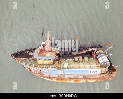Aus der Vogelperspektive eines rostigen verlassenen Fischerbootes mit dem Kennzeichen S 226, das in flachem, trübem Wasser ruht und Algen auf dem Deck sichtbar ist. Stockfoto
