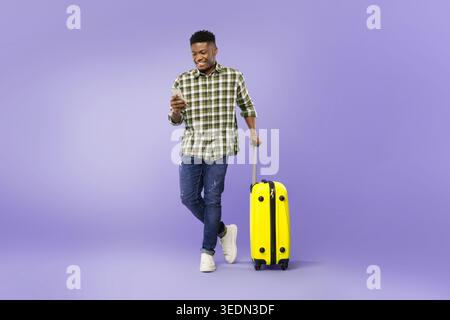 Mann mit gelbem Koffer überprüft das Telefon in einer Reiseeinstellung vor violettem Hintergrund Stockfoto
