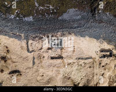 Luftaufnahme der Ruinen eines verlassenen Bauernhauses auf sandigem Gelände in der Nähe einer dunklen felsigen Küste in Hafnarfjordur, Hafnarfjardarkaupstad, Island. Stockfoto