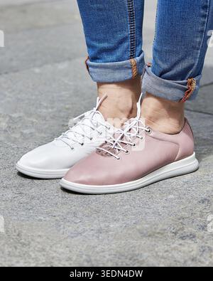 Close-up Street-Style aus aufgerolltem Denim und nicht passenden Sneakern aus weißem und weichem Leder zum Schnüren auf einem Bürgersteig aus Beton. Stockfoto