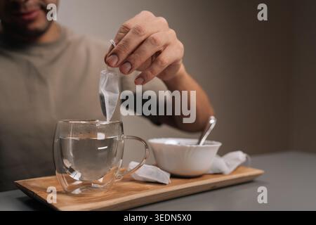 Nahaufnahme einer Person, die wohltuenden Kräutertee aufbrüht, Teebeutel in einen transparenten Becher mit dampfendem Wasser auf einem Holztablett senkt, wodurch ein beruhigendes Getränk entsteht Stockfoto