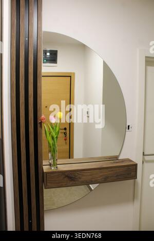 Sauberer und moderner Eingangsbereich mit einem hölzernen Konsolenregal, dekorativem Spiegel und einer Vase mit Tulpen. Stockfoto