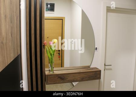 Minimalistischer Flur mit Holztür, rundem Spiegel und Tulpen in Vase auf Konsolentisch, die einen warmen, modernen Hauseingang schaffen. Stockfoto