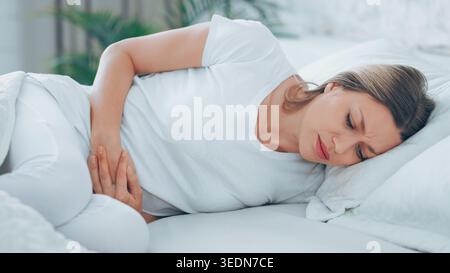 Die Frau ruht sich mitten am Tag auf dem Bett aus, während sie den Magen in Unwohlsein hält Stockfoto