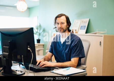 Porträt eines männlichen Rezeptionisten, der am Schreibtisch mit Computer im Krankenhaus sitzt Stockfoto