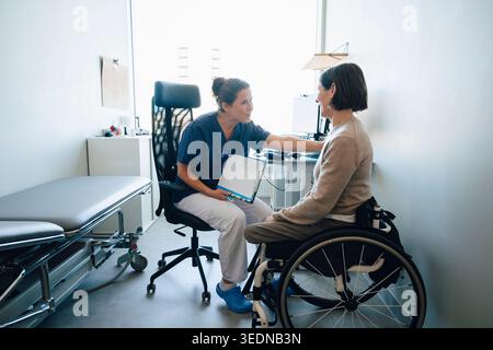 Glückliche weibliche medizinische Fachkraft, die positive medizinische Aufzeichnungen zu Frau, die auf Rollstuhl im Untersuchungsraum sitzt, teilt Stockfoto