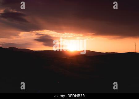 Dramatischer Sonnenuntergang über sanften Hügeln mit glühendem Himmel und Wolken, malerische Landschaft mit warmem orangenem Licht und Silhouetten, friedliche abendliche Natur Stockfoto