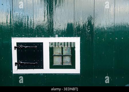 Authentisch verwitterte grüne Holzwand mit einem traditionellen weißen Fensterrahmen und selektivem Fokus. Verfügt über einen schwarzen Verschluss mit Eisenscharnieren und einem s Stockfoto