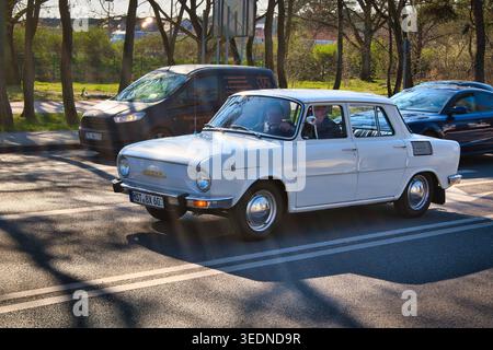 Oldtimer in weiß ŠKODA 100, Fahrt auf der sonnendurchfluteten Stadtstraße mit Verkehr und Bäumen - die Küstenstadt Świnoujście. Stockfoto