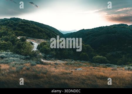 Malerische Berglandschaft mit gewundenen Feldwegen durch grüne Hügel und Wald unter dramatischem Himmel bei Sonnenuntergang, Panoramablick auf die Natur ideal für Reisen, Stockfoto