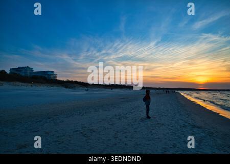 Einsame Figur, die den farbenfrohen Sonnenuntergang über Sandstrand und die ruhige Küste des Ozeans mit weit entfernten Gebäuden beobachtet - die Küstenstadt Świnoujście. Stockfoto