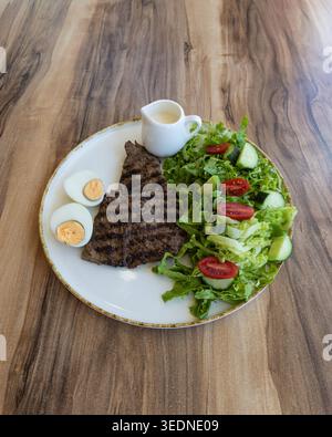 Blick von oben auf ein gegrilltes Rindersteak, serviert mit frischem grünem Salat, Kirschtomaten, Gurke und gekochten Eiern auf einem rustikalen Holztisch. Gesund ausgewogen Stockfoto