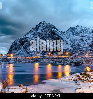 Winterlandschaft in der Dämmerung in nordischer Umgebung mit teilweise gefrorenem See, der Dorflichter und Straßenlaternen reflektiert. Schneebedeckte Berge Stockfoto