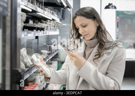 Eine Frau in einem neutralen beigefarbenen Mantel verwendet ihr Smartphone, um Informationen über ein verpacktes lokales Produkt in einem modernen Bauernladen zu scannen oder zu lesen. Kühlte sie Stockfoto