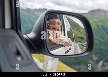 Junge Frau, die sich in einem Auto-Seitenspiegel auf einer Reise in die Alpen reflektiert und Freiheit und Reisen annimmt Stockfoto