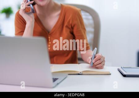 Geschäftsfrauen sprechen Smartphone schreiben Notizen Planen Sie Aufgaben vom Home Office Schreibtisch Laptop Computer ferngesteuerte Hybrid-Arbeit Stockfoto