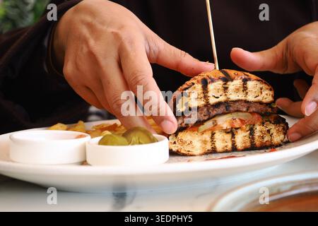 Hände halten einen Burger mit Belag auf einem weißen Teller Stockfoto